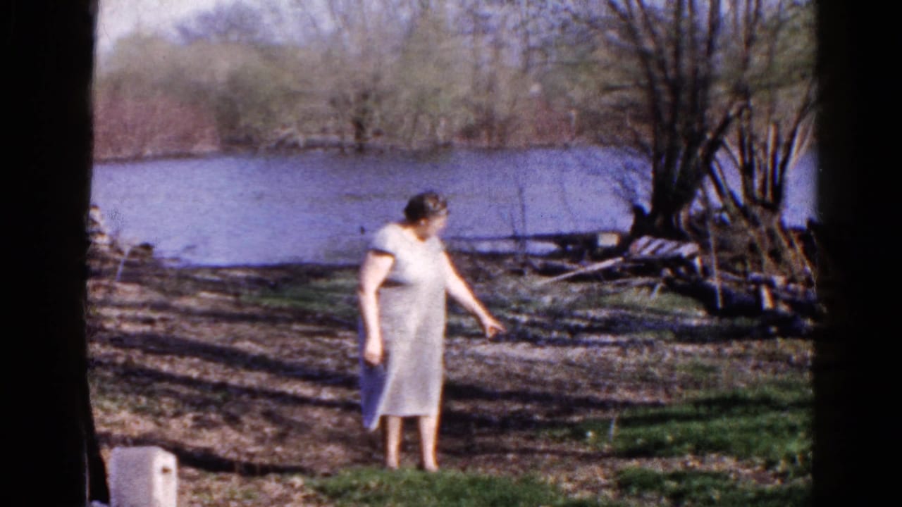 In 1961 Wauconda, Illinois, an adult woman in a light dress stands barefoot near a wooded riverbank.