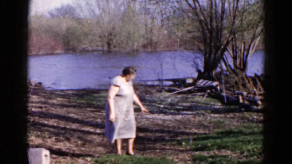 Adult woman in a light dress looks toward the ground on a wooded riverside in Wauconda, Illinois, 1961.