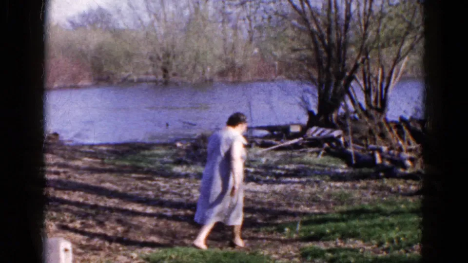 Adult woman walks barefoot across a shaded riverbank, with water and early-season trees in the background.