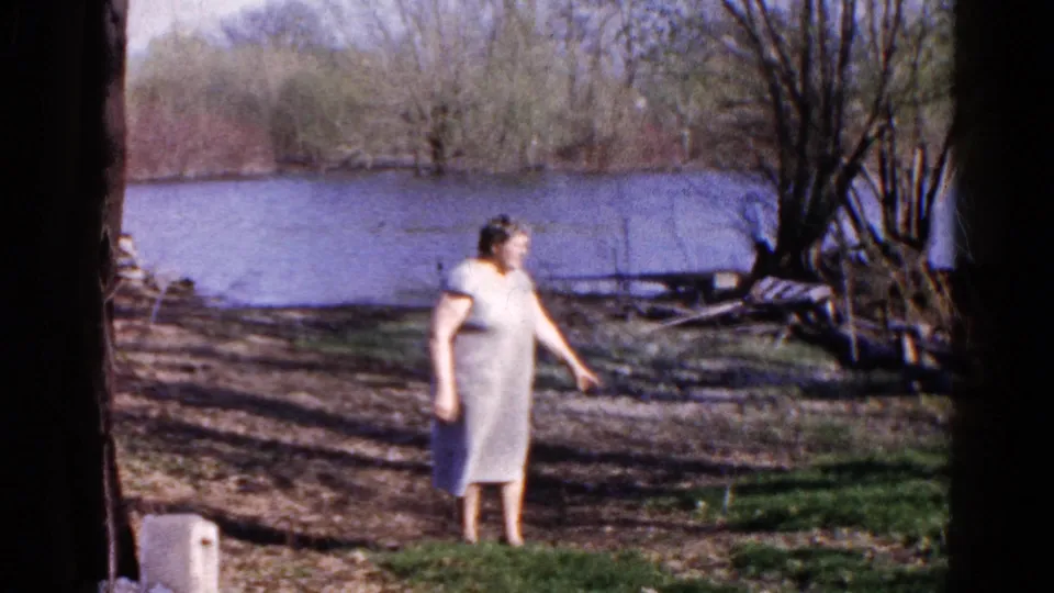Adult woman in a light dress stands on a Wauconda lakeshore in 1961, pointing near the shoreline as trees and water fill the background.