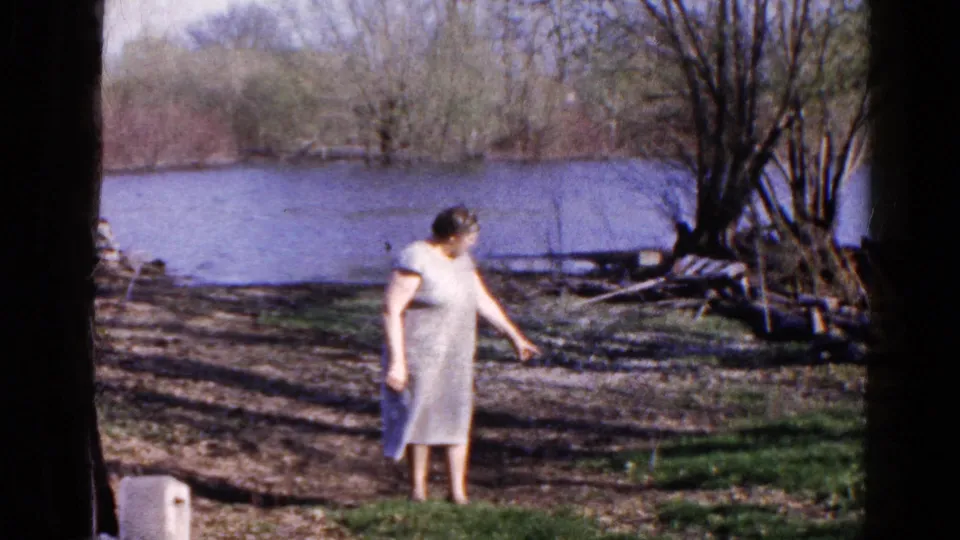 Adult woman continues pointing by the Wauconda lakeshore, framed by a tree trunk, bare ground, brush, and blue water in 8mm footage.
