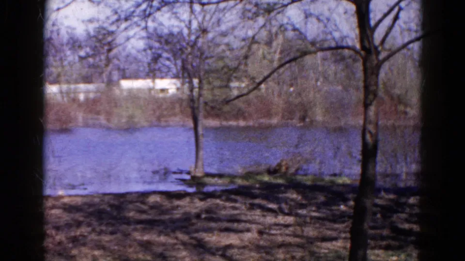 Wauconda, Illinois lakeside frame from 1961 showing bare branches, calm water, and a pale building beyond the far bank.