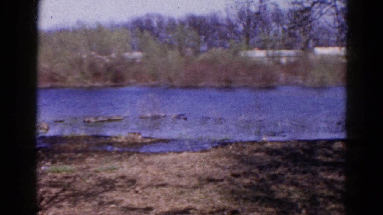 1961 Wauconda 8mm frame showing a river, bare shoreline, brush, trees, and distant pale buildings across the water.
