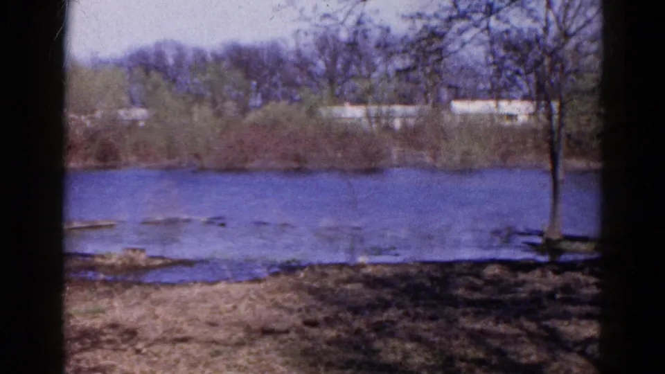 1961 Wauconda home-movie frame of blue river water, bare shoreline trees, brush, and low light structures beyond the far bank.