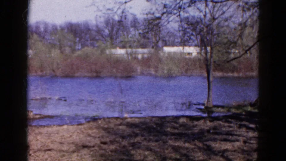 1961 Wauconda 8mm frame with a quiet river, muddy foreground, leafless trees, brush, and distant pale buildings across the water.