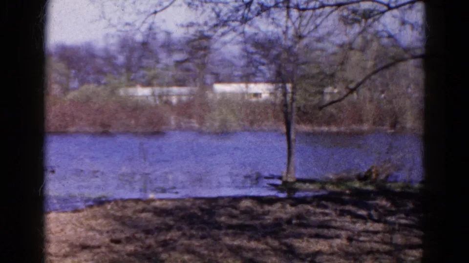 1961 Wauconda home-movie frame showing blue river water, a bare tree at the bank, brush, and pale distant buildings.