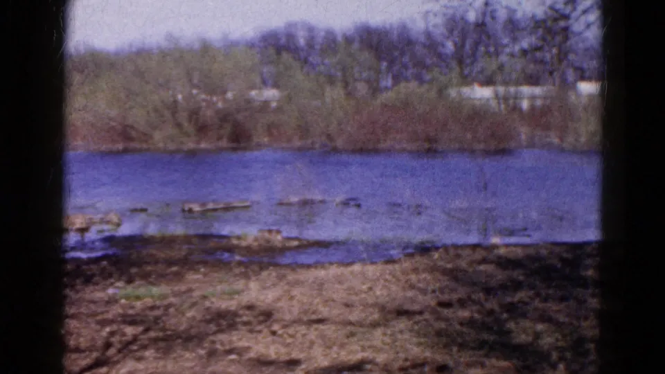 1961 Wauconda, Illinois frame showing calm water, shoreline logs, muddy bank, and a distant treeline with pale buildings partly visible.
