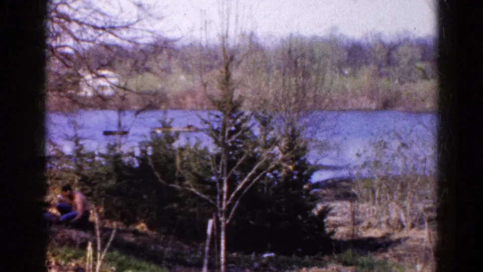 1961 Wauconda, Illinois home-movie frame of an adult sitting by the river, partly framed by shrubs and bare branches.