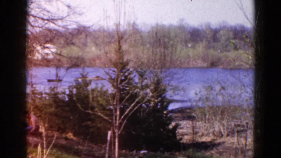 1961 Wauconda, Illinois 8mm frame with a partly cropped seated adult at lower left and a calm river behind shrubs and trees.