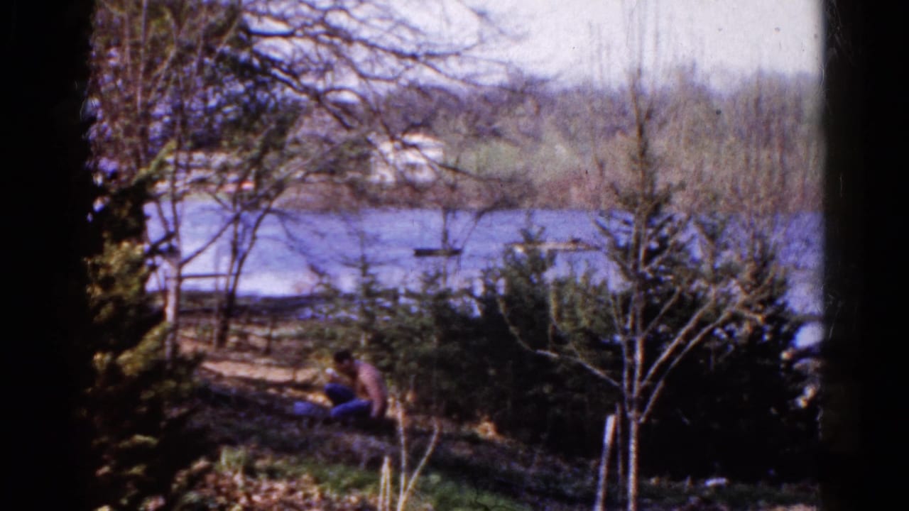 Man seated on a grassy slope near a lake with trees and shrubs around him.