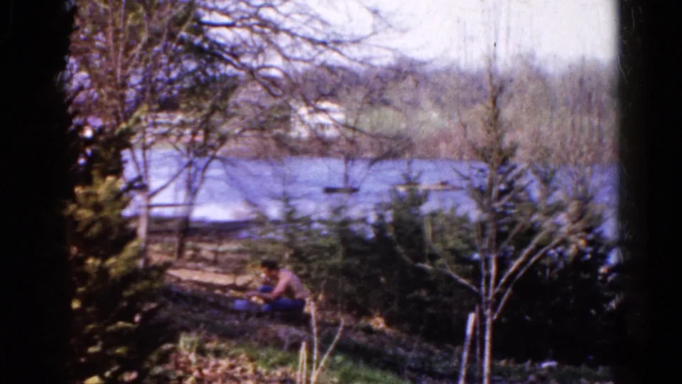 The 1961 Wauconda home movie shows a seated man low in frame, with calm water, bare branches, and a lakeside fence behind him.