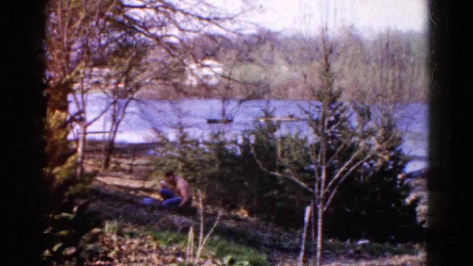 A man remains seated on the Wauconda lakeside slope, surrounded by shrubs and bare trees, with blue water and distant shore visible behind him.