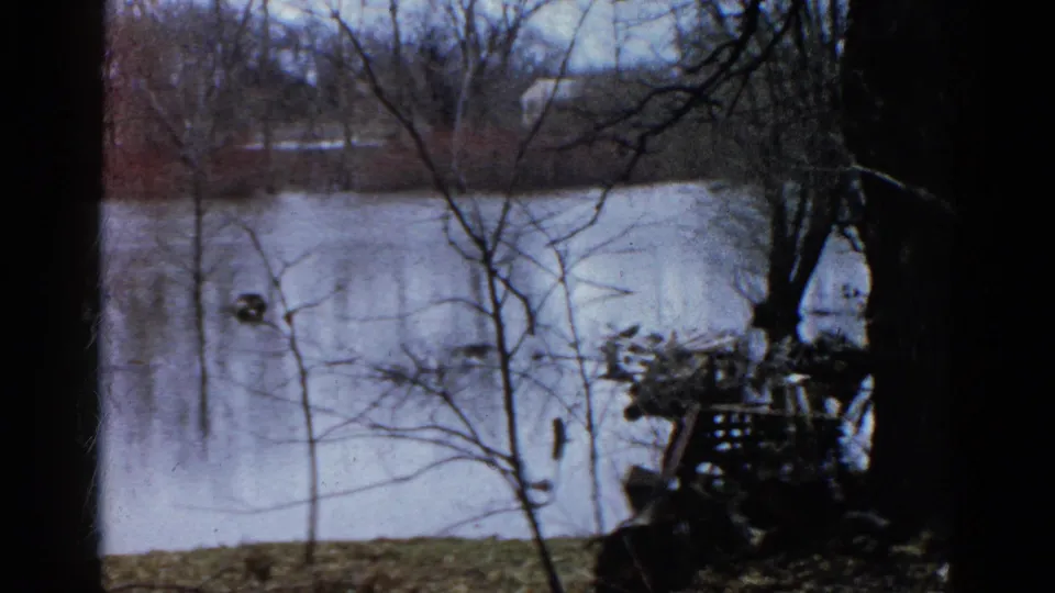 Soft 1961 Wauconda 8mm frame of a pond through winter branches, with a dark bank-side form at right.