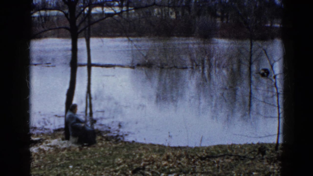 1961 Wauconda, Illinois 8mm frame showing a seated person near a quiet lake, framed by bare trees and a grassy winter bank.