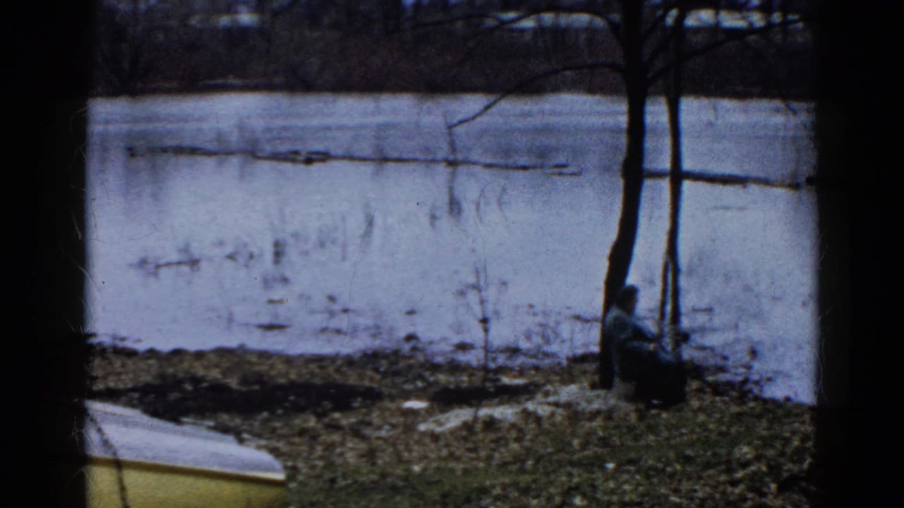 A person sits near a tree on the shore of a partially frozen lake in 1961 Wauconda, Illinois home-movie footage.