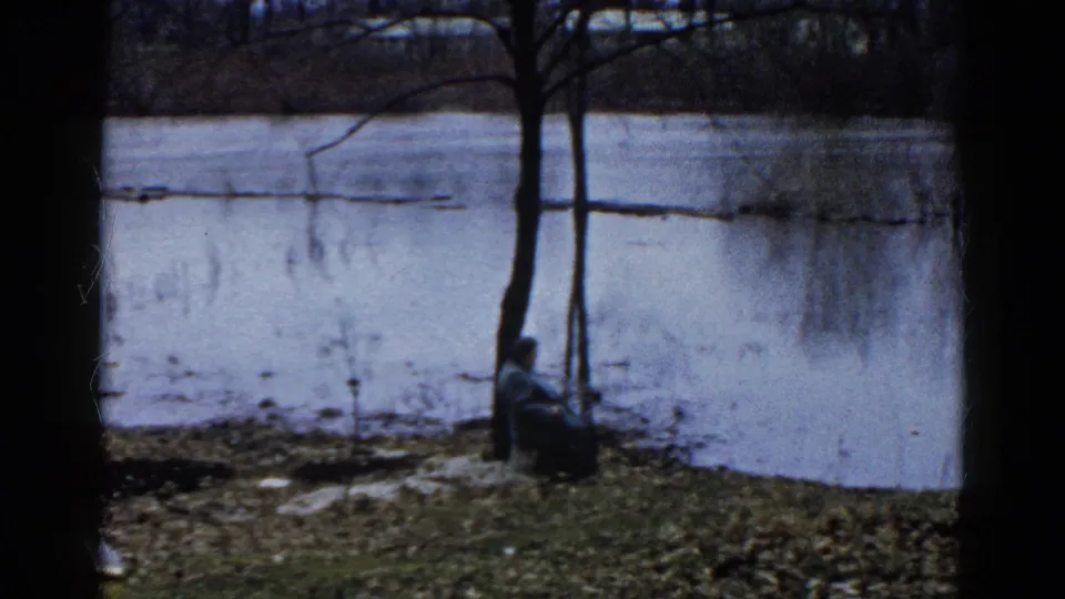 1961 Wauconda lakeshore frame with a seated figure at a bare tree, winter grass, and pale frozen water behind.