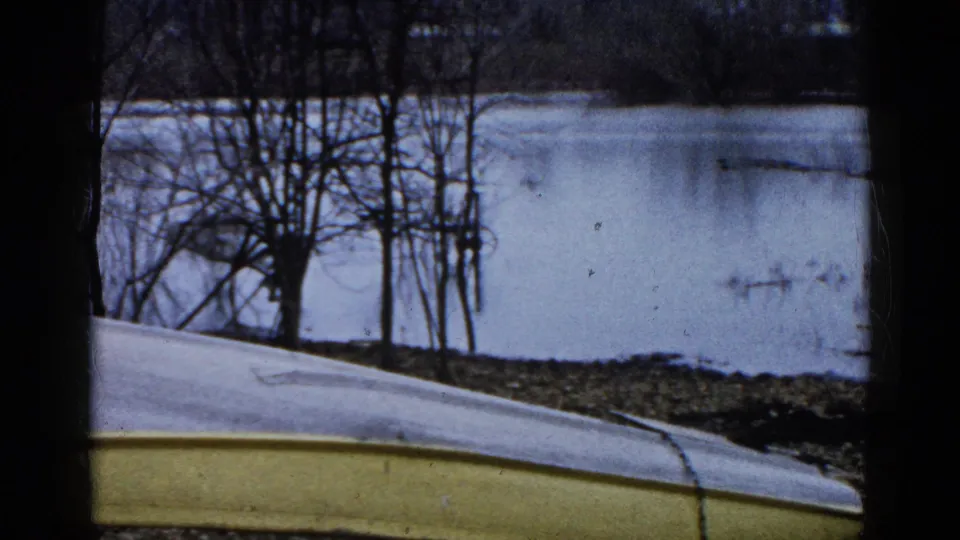 1961 Wauconda, Illinois home-movie frame of a flipped boat near icy water, with leafless trees and muted winter reflections in the background.