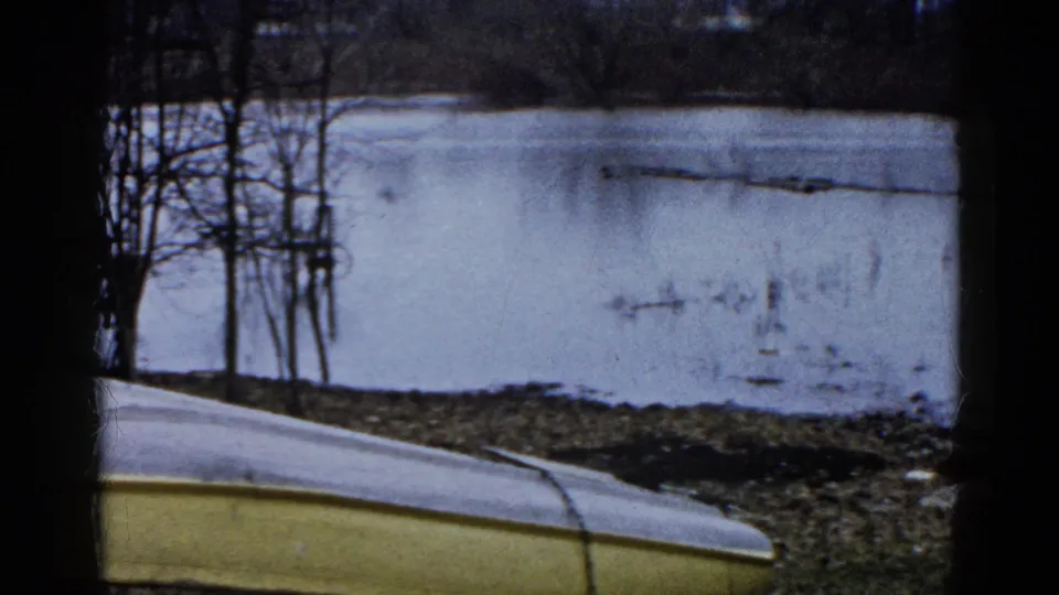 1961 Wauconda, Illinois 8mm still showing the lower edge of an overturned boat, muddy winter shore, and pale icy lake beyond.