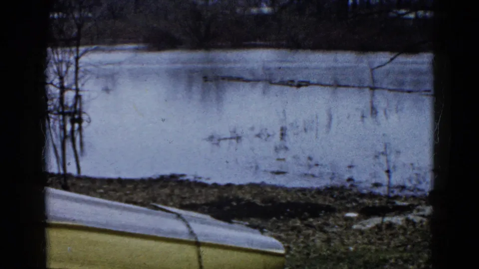 1961 Wauconda, Illinois winter lakeshore frame with an overturned yellow boat edge, muddy bank, icy water, and distant bare trees.