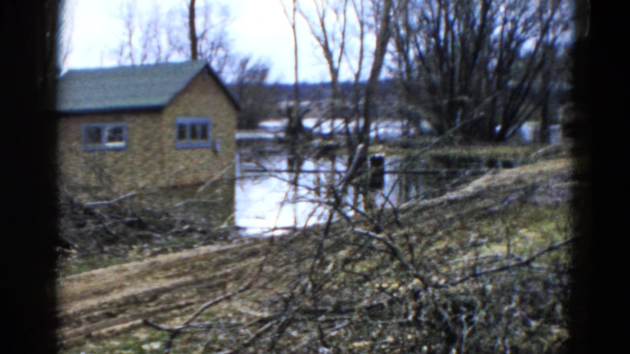 1961 Wauconda, Illinois 8mm frame showing a small brown lakeside building, still water, bare trees, brush, and a dirt path.