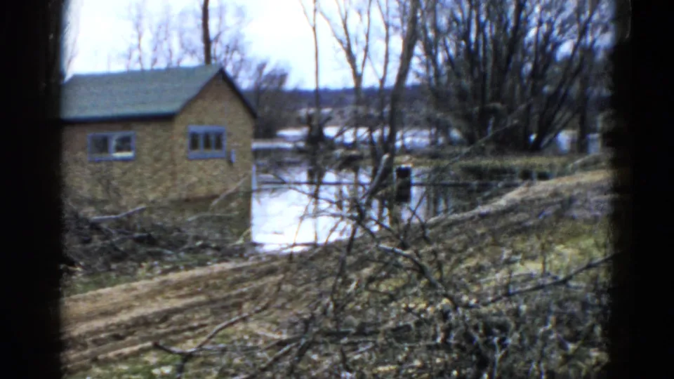 1961 Wauconda, Illinois 8mm frame showing a small brown lakeside building, still water, bare trees, brush, and a dirt path.