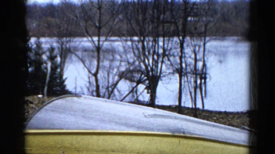 1961 Wauconda, Illinois lakeside frame with still water, bare trees, shoreline brush, and a pale curved roof or boat edge in the foreground.