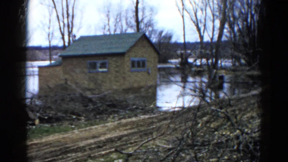 Home-movie view from Wauconda in 1961 showing a flooded brick house framed by dark edges and bare trees.