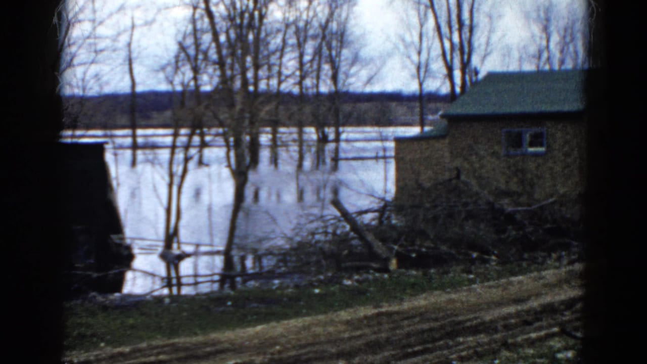 Small building beside floodwater with bare trees and muddy ground.