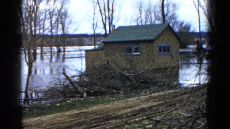 1961 Wauconda, Illinois home-movie frame of a small building beside floodwater, with bare trees, brush, and muddy ground in view.