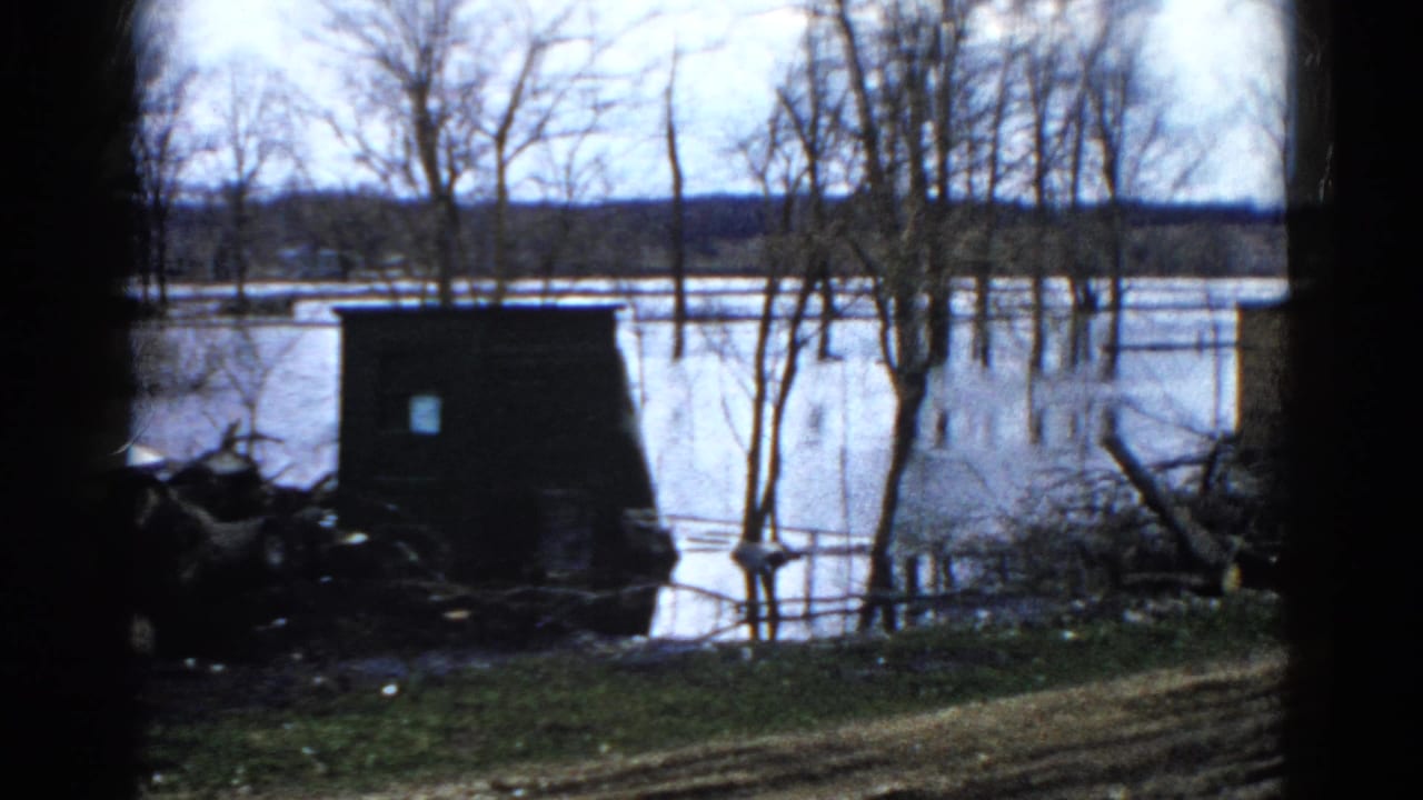 1961 Wauconda 8mm frame showing floodwater around bare trees near a small dark structure and distant wooded horizon.