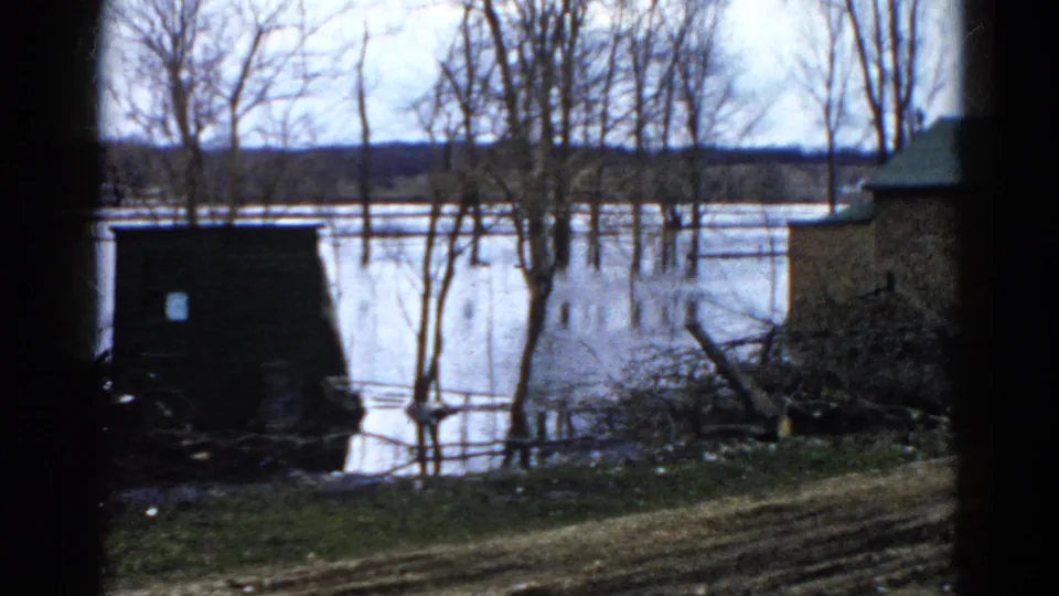 1961 Wauconda home-movie frame with floodwater, leafless trees, a dark shed-like structure, brush, and a house edge.