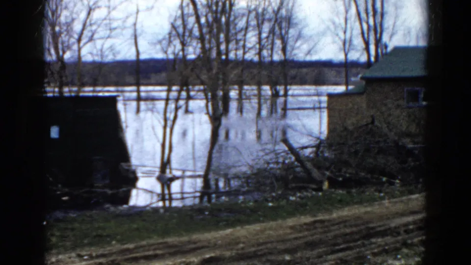 1961 Wauconda frame showing a small home and dark structure beside floodwater with bare trees and brush in view.