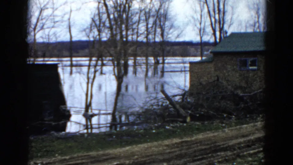 1961 Wauconda 8mm frame of floodwater near a small house, bare trees, brush, and a distant wooded horizon.