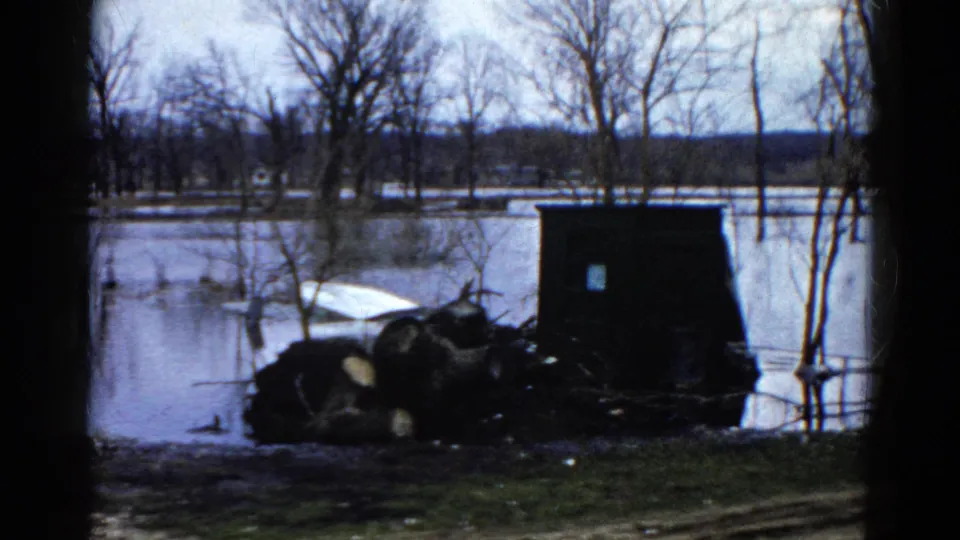 1961 Wauconda, Illinois frame with a dark shed-like structure, piled logs, debris, and floodwater among leafless trees.