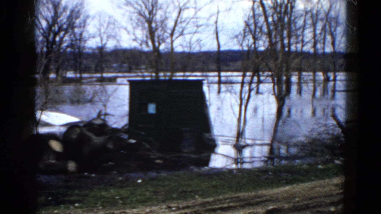 1961 Wauconda, Illinois frame showing a dark shoreline structure, piled logs, and floodwater extending through leafless trees.