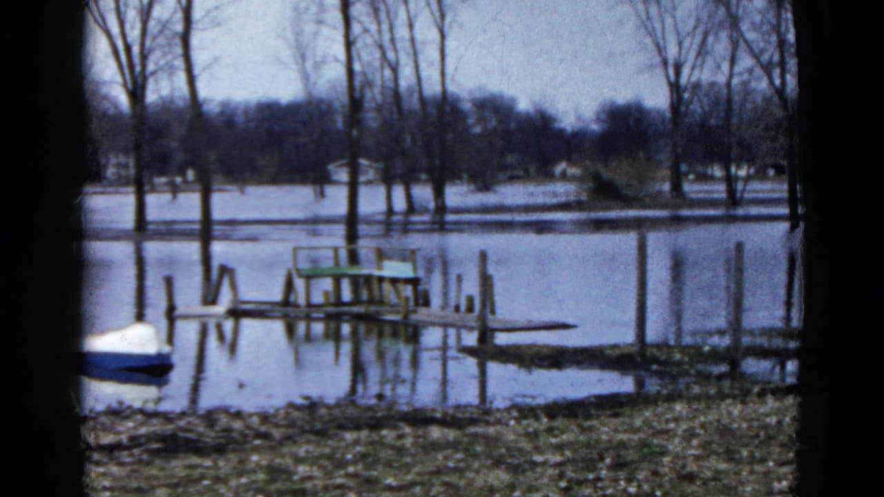 1961 Wauconda, Illinois 8mm frame showing a partially submerged dock, green bench, calm lake water, bare trees, and a small boat at left.