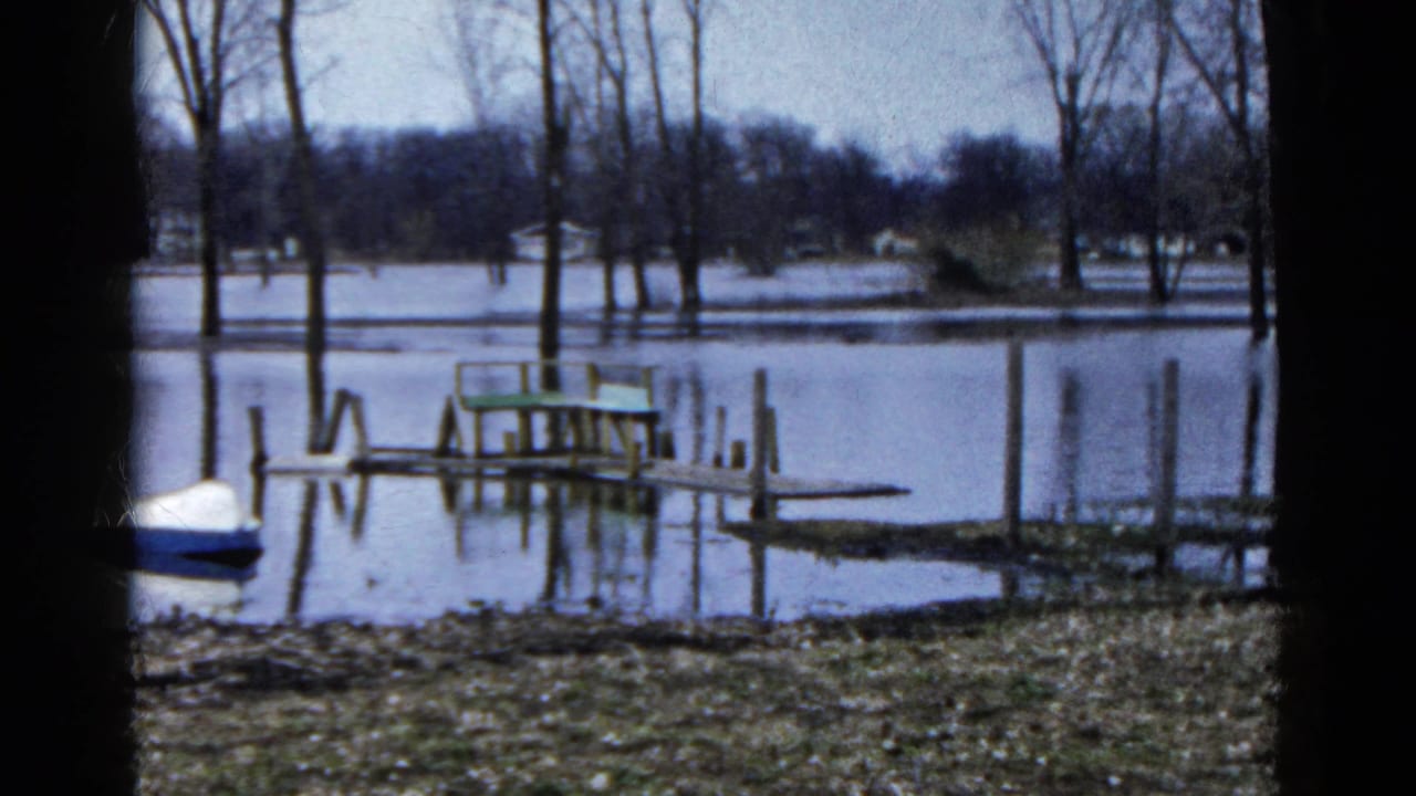 1961 Wauconda, Illinois home-movie frame of a flooded dock and green bench near a small boat, with bare trees and distant shoreline beyond.