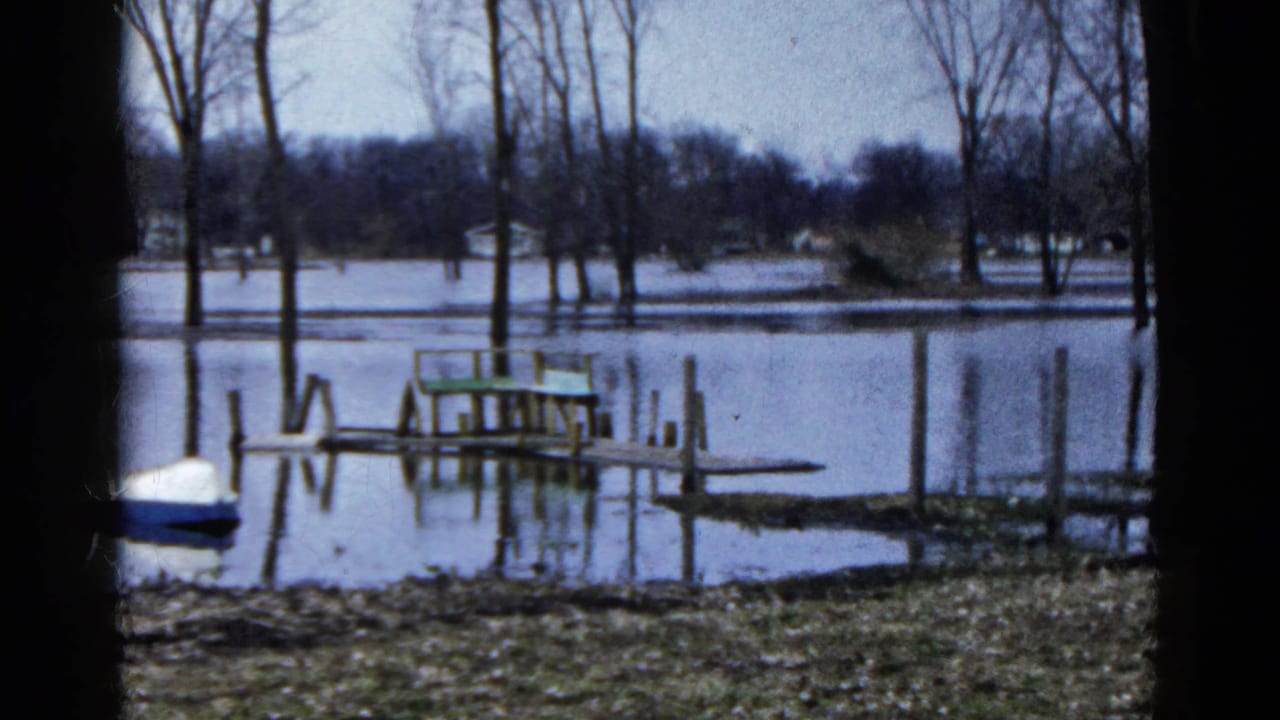 1961 Wauconda, Illinois 8mm frame showing high water around dock posts, a green bench, bare trees, shoreline grass, and a small boat at left.
