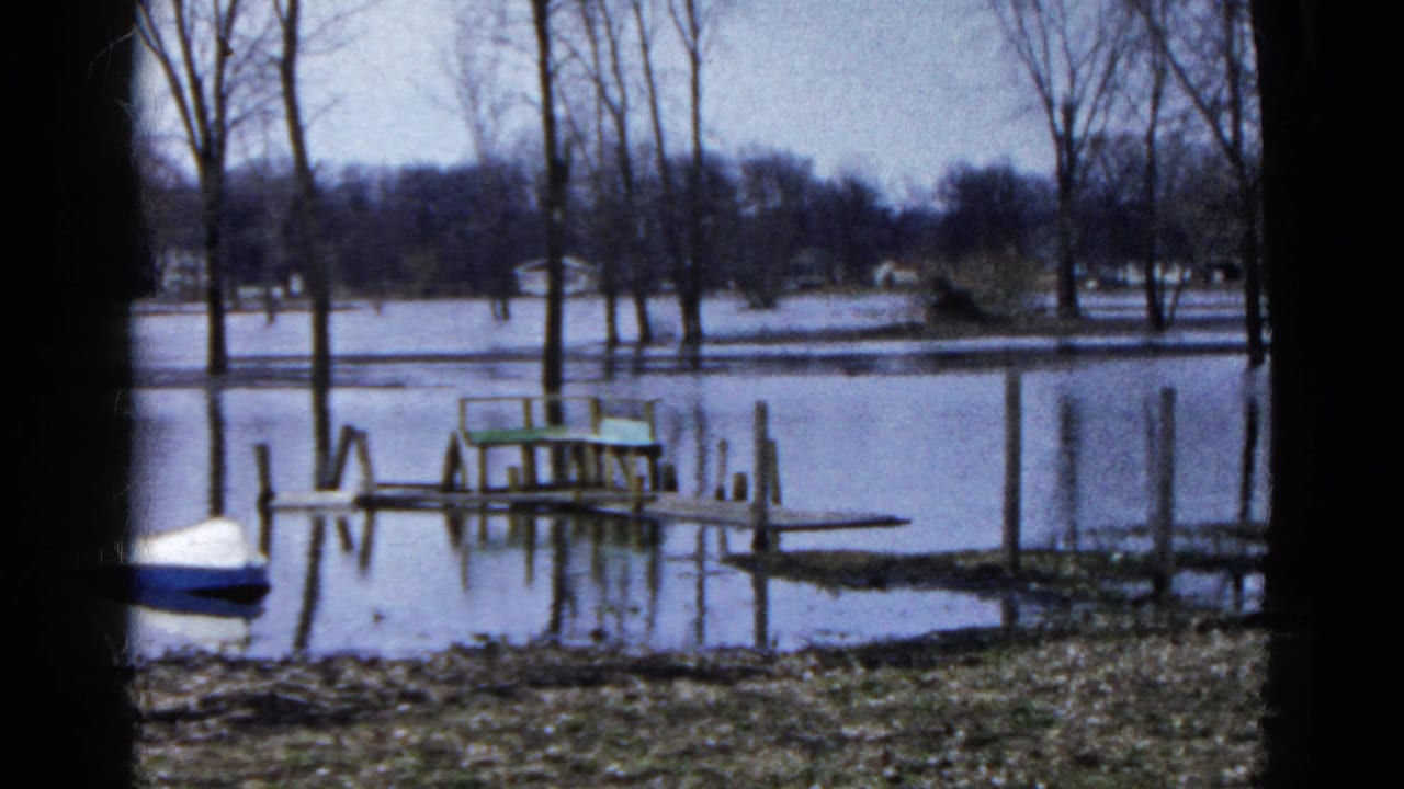 1961 Wauconda, Illinois frame of a flooded lakeshore, partly submerged dock, bare trees, and a blue-and-white boat near the bank.