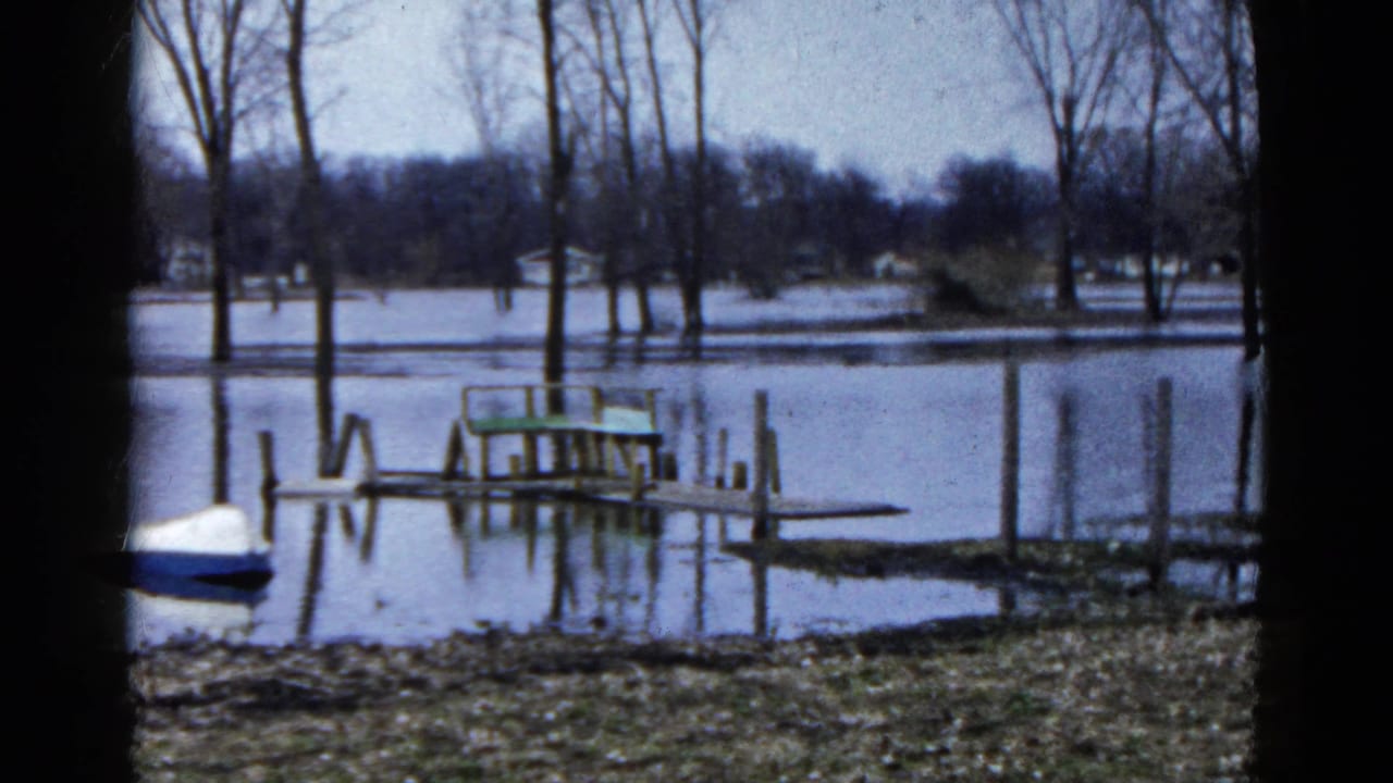 1961 Wauconda, Illinois lakeside frame showing floodwater over the shore, a low dock, vertical posts, and a small blue-and-white boat.