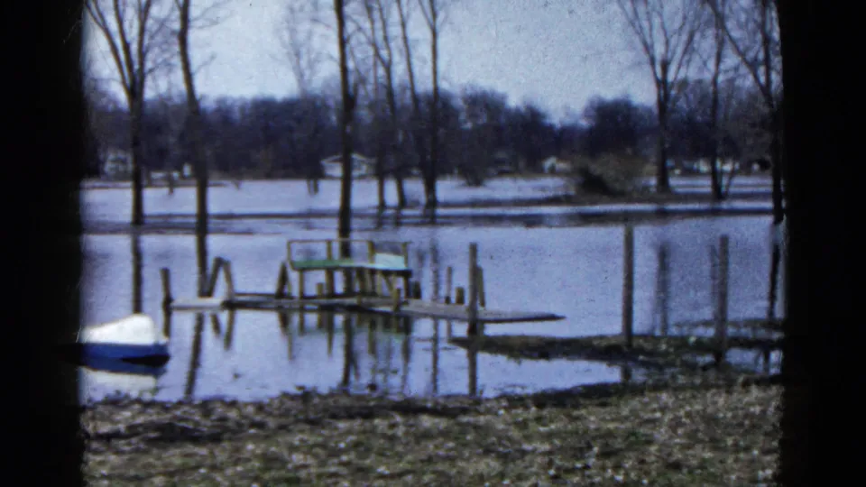 1961 Wauconda, Illinois lakeside frame showing floodwater over the shore, a low dock, vertical posts, and a small blue-and-white boat.