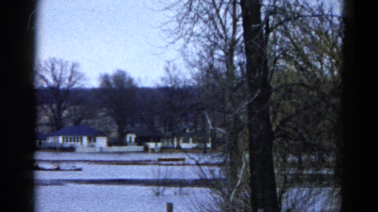 1961 Wauconda, Illinois 8mm frame showing a snow-covered exterior view with bare trees, distant low buildings, and dark frame edges.