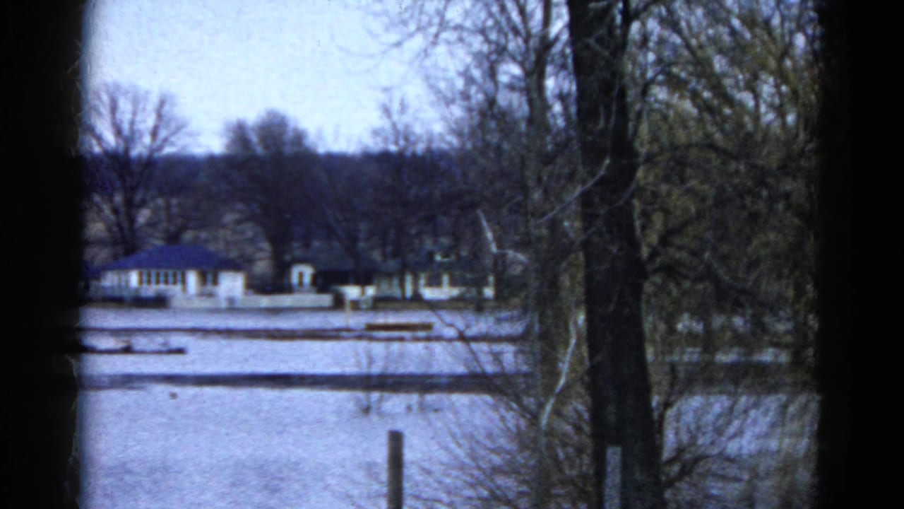 1961 Wauconda, Illinois frame showing a quiet snowy exterior with tree trunks, bare branches, and buildings in the distance.