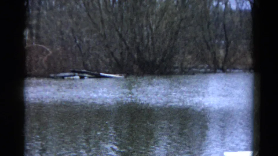 1961 Wauconda, Illinois home-movie frame with blue-gray floodwater, bare trees, and scattered debris near the wooded shoreline.