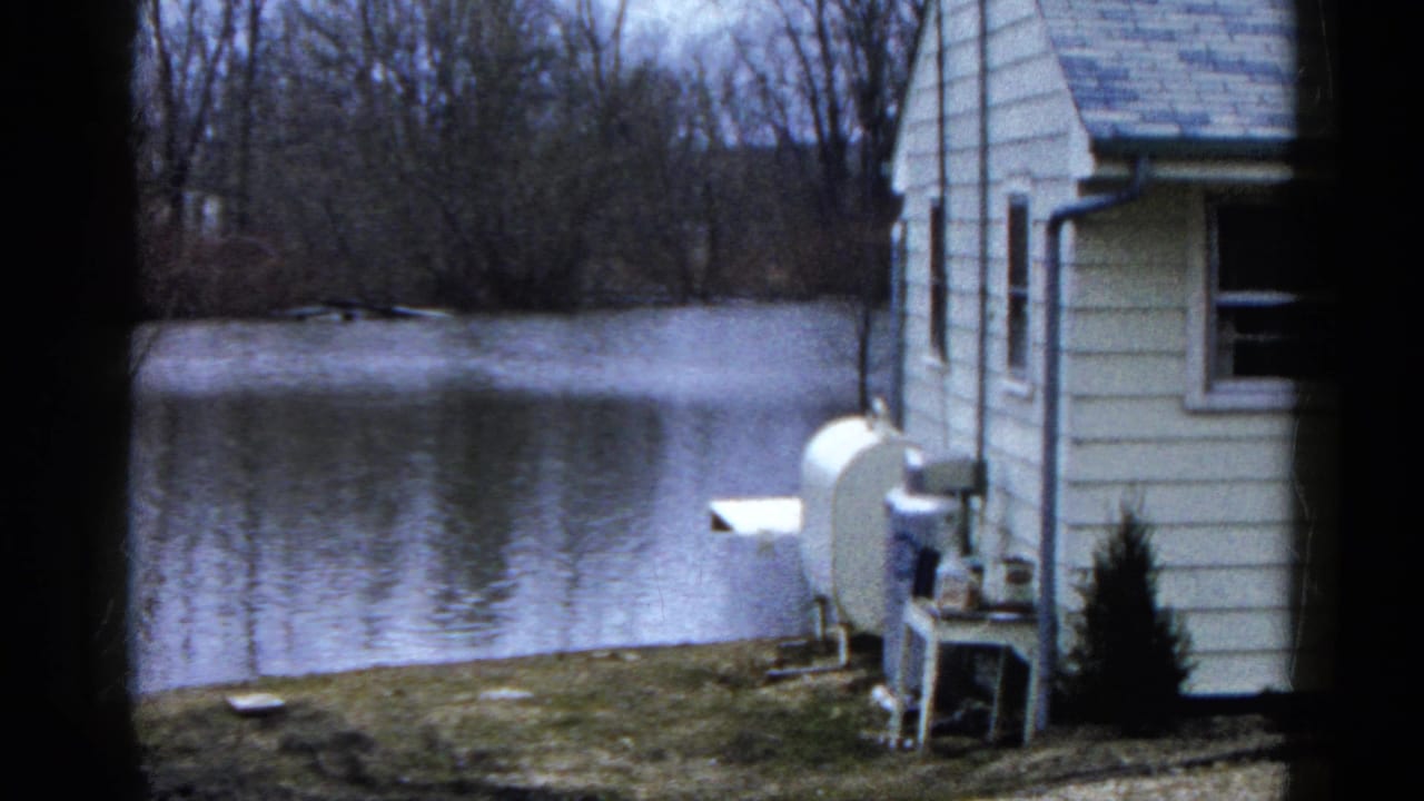 White-sided lakeside house with yard equipment beside calm water and bare trees.