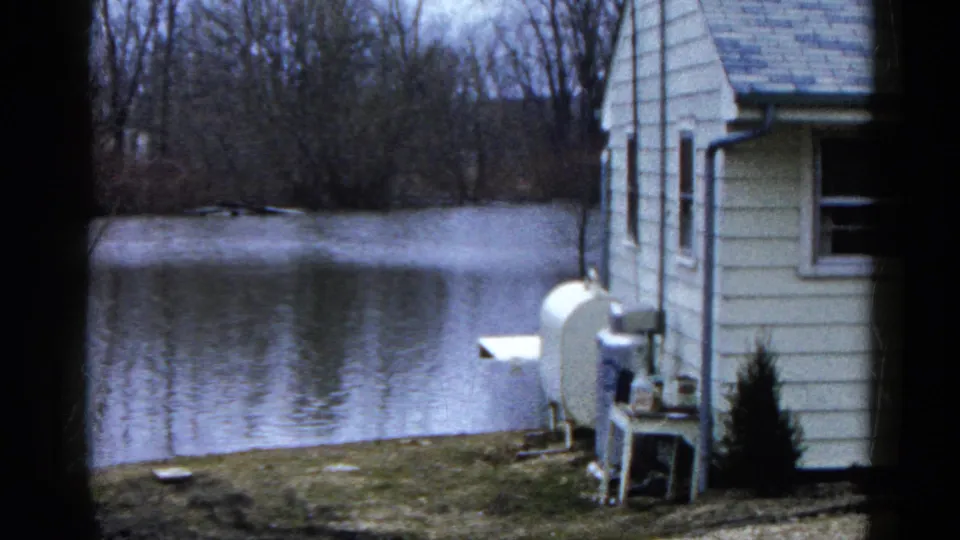 1961 Wauconda, Illinois 8mm frame showing a white-sided house edge, outdoor equipment, lawn, lake, and bare trees across the shore.