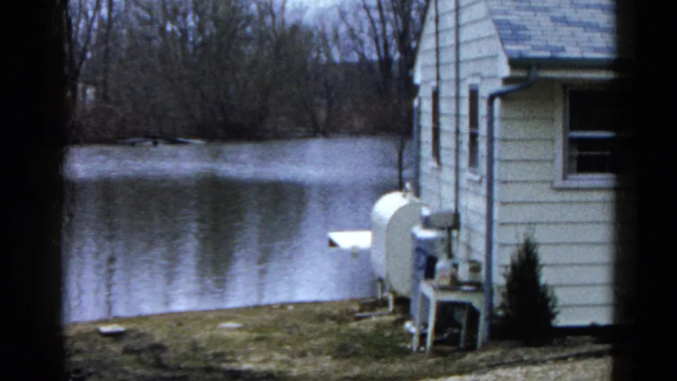 The lakeside house remains at frame right, with yard equipment near the wall and reflective water stretching toward bare trees.
