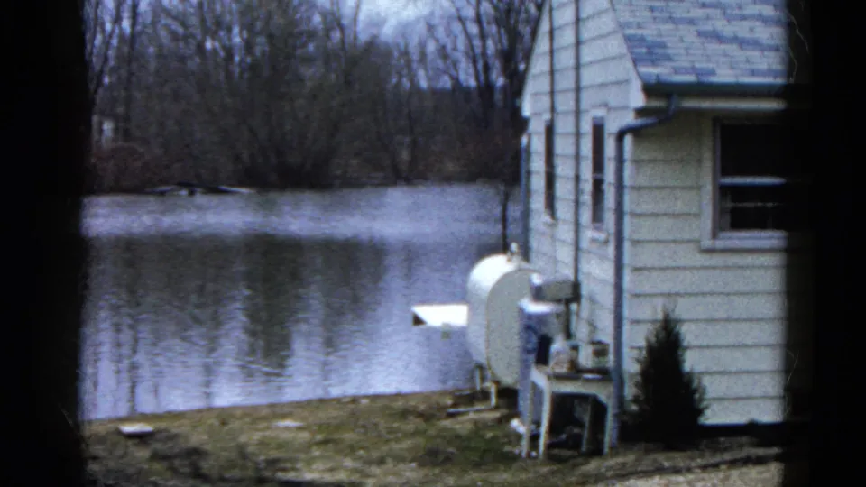 Final frame holds the same lakeside residential view, with white siding, outdoor equipment, calm water, and leafless trees across the lake.