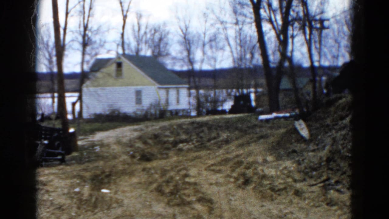 1961 Wauconda home-movie frame showing a white house with yellow trim near a lake, seen past a dirt path and bare trees.