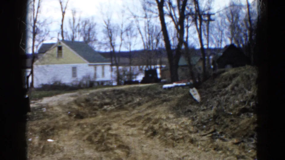 The lakeside house remains framed by bare trees, with the dirt path and grassy bank leading toward the water.